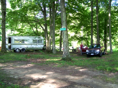 A camper and truck at a wooded riverside campsite under lush hardwood trees
